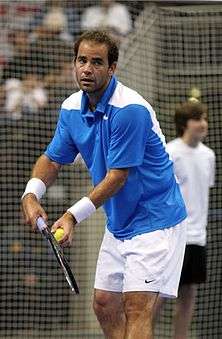 A man, with a modern racket in his right hand and a tennis ball in his left hand, prepares to serve
