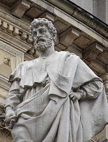 View looking up at a white marble statue of a man with a curly beard wearing classical robes