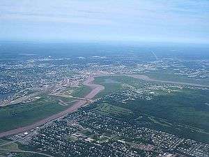 An overhead view shows a river winding through the city of Moncton. There is parkland and a golf course near the water, and city streets further out.