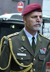 An older man with a gray beard, red beret, and olive green military suit.