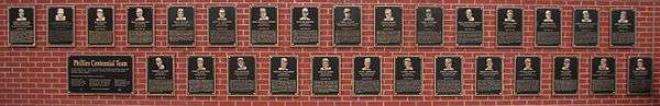 Forty black metal plaques are mounted on a brick wall. They are inscribed with gold images of human faces and text.
