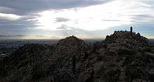 a view of the craggy tops of two small mountain peaks in the Phoenix Mountain preserve, with the city of Phoenix in the background.