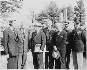 Seven men in suits and one in an Army uniform in a car park.