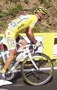 A cyclist in a yellow and white jersey with black trim rides alongside a yellow guard rail.