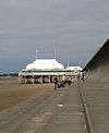 Short pier above sand, surmounted by white pavilion with flag poles.