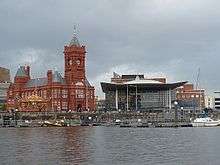 Red brick Victorian style building with clock tower and the Senedd, with water in foreground
