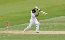 A man wearing a helmet and white clothes, in the motion of swinging a cricket bat.