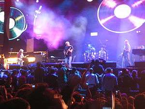 A concert stage lit by purple lighting. Four men are performing on the stage as a crowd stands in front of it. Behind the men are video screens displaying images of vinyl records.