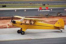 A bright yellow, single-propeller light airplane is sitting on a paved surface at what appears to be an airport. A man is sitting in the pilot's seat, and the propeller is turning. On the tail of the plane is a logo consisting of a small bear holding a sign that says, "Cub". The number on the tail rudder is N70843.