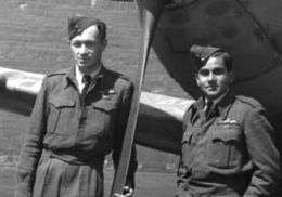 Two men in air force uniforms stand in front of a fighter aircraft