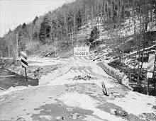 Black and white photograph looking along the roadway of a bridge flanked by low stone walls with a large "ROAD CLOSED" sign on the bridge. Snow covers some of the bridge and the forested hillside in the background.