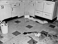 A black and white image of a checkered tiled floor with a right-angled row of light-colored cabinets at the rear. On the floor in the foreground are several large dark smears, with a small piece of paper on the ground nearby. At the left is a small white plastic bucket flled to over the top with trash; some cans and a telephone handset are visible. In the rear, near the cabinets, is a roll of paper with part of it spilling off to the right; underneath it are what appear to be two books