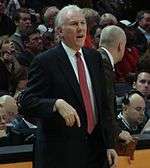 A man, wearing a black suit, white shirt and red tie, is standing in front of the spectators.