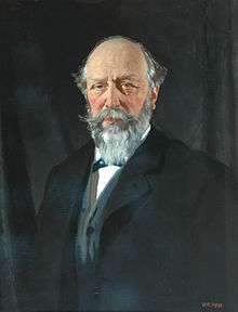 Portrait of a balding middle-age man with grey hair and beard, wearing a dark overcoat over a blue jacket and white shirt.