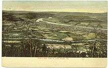 A tinted postcard depicts a town as seen from a hillside. It is laid out on a triangle of flat land between two converging streams. The larger and more distant of the two streams is flowing from mountains, while the smaller stream flows through farmland.