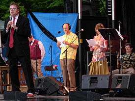full length portrait of Garrison Keillor with a microphone and three performers behind him and someone seated on the stage