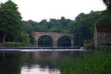 A stone two-arched bridge across a river, viewed along the river, both ends hidden by trees. A weir is in front of the bridge, at the right end of which is a two storey building.