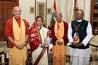 A color photo of the President flanked by Hare Krishna followers in robes, holding a copy of the Bhagavad Gita in Russian.