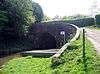 The canal enters the tunnel through and arch and on the right a road curves towards the left to cross the bridge.