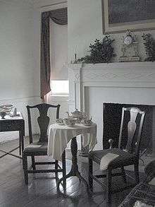 Photograph of a bedroom, showing two chairs and a tea table in front of a fireplace. The room is painted white and the furniture is of a dark wood.