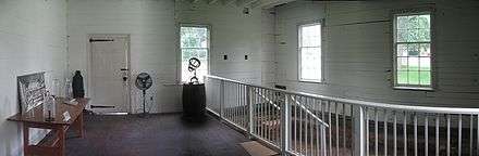  White pained interior of a room with wooden walls, three windows, and door. There is a wooden table at left and a white railing at left.