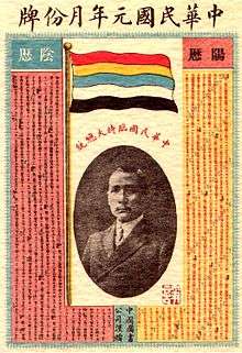 A calendar with a picture of a Chinese man in the center. On top of it stands a flag with five horizontal stripes (red, yellow, blue, white, and black).