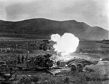 Several artillery guns fire in unison in a field