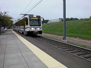 A white and yellow light rail train approaches an empty station platform with overhead lines visible above.