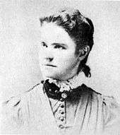 A washed-out monochrome photograph portrait of a young woman with shoulders squared to the camera, the head turned to the left, a neutral-colored dress topped by a dark ruff and white lace, the woman's dark hair pulled back behind her ears to fall down below her collar