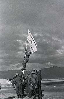 Israeli soldiers stabilize a flag pole whilst another soldier climbs it in order to raise an improvised flag; the soldier is seen about halfway up the flag pole. Other soldiers look on.