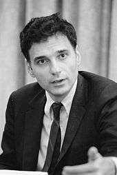 Young-looking Nader at 40+ years old gesturing as he speaks, wearing a coat and tie with unruly wavy dark hair.