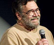 Head and shoulders of a bearded older man in glasses. Smiling, with eyes nearly closed, he is wearing a plain sweater and holding a microphone.