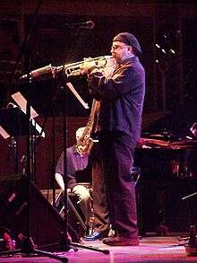 A man on a stage wearing all black and a cap on his head, playing a trumpet into a microphone. Behind him is a man holding a saxophone and another man sitting in a chair. Music stands and additional microphone stands are on the stage in front of them.