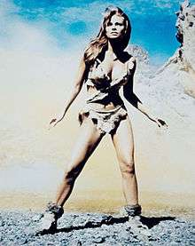 Welch in fur bikini, in smoky, rocky surroundings, stands with feet braced apart, hands away from sides, tensed as if seeing a threat in the distance.