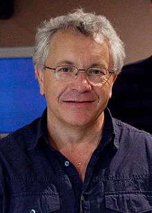 A white haired man is seen wearing a blue collared shirt.