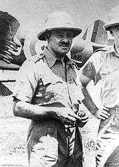 Portrait of a man in tropical military uniform. He is wearing a bucket hat, and military aircraft can be seen in the background.