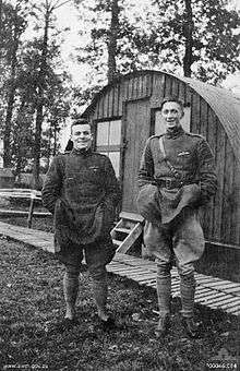 Two men in military uniform standing side-by-side. The man on the left if short and stocky, while the man on the right is tall and thin. In the background is a cabin.