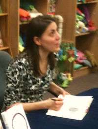 Stead, a black haired woman in her 40s, sits at a table and signs a book while looking slightly upward. Behind her are wooden shelves filled with colorful toys.