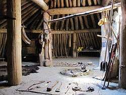 Earth lodge interior showing the central wooden pillars, wooden walls, a packed clay floor, a fire pit, back rests, a table with food, a fur rug, cattail mats, various other furnishings, and a canopy bed