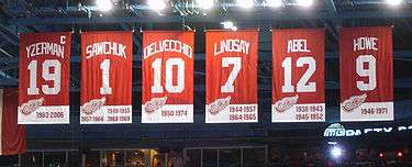 A row of six red banners.  The banners, from left to right, read "Yzerman 19" "Sawchuk 1" "Delvecchio 10" "Lindsay 7" "Abel 12" "Howe 9". The Yzerman banner has a small "C" at the top right corner.