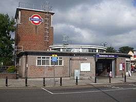 A red-bricked building with a rectangular, dark blue sign reading "REDBRIDGE STATION" in white letters all under a dark blue sky with white clouds
