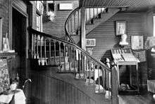 A black-and-white image of the curving wooden staircase with Gothic-arched banister described in the accompanying text. There are many artifacts, paintings and other decorations on the walls