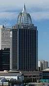 Ground-level view of a 30-story building with a square cross section and a dark glass facade; on the roof there is large, white, latticework structure that tapers into a spire.