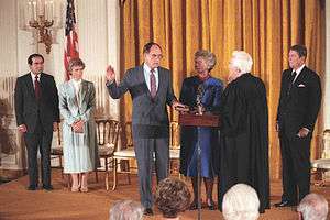 An elderly white haired man in judicial robes swears in a middle-aged man in a suit as several people look on.