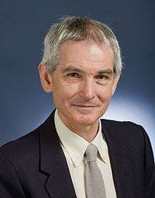 Official photograph of middle aged man with grey hair and no other facial hair, wearing a suit, smiling into the camera.