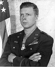 Head and shoulders of a stern-faced white man with his arms folded over his military jacket, standing in front of a wall map and an American flag. Rows of ribbon bars are on his left breast, pins adorn his lapels, and a star-shaped medal hangs from a ribbon around his neck.
