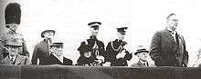News photo of Reid, making a speech, outdoors in a reviewing stand. A number of other dignatories are seated in the stand, and behind them stand four members of the armed services, in dress uniform.
