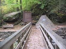 Photo taken from a wooden footbridge with handrails over a rocky creek in a green forest. At the end of the bridge are a large boulder with a bench below it to the left, a trail sign labeled "Waters Meet" and "The Falls Trail" above a map of the trail in the center, and a natural stone monument with a metal plaque to the right.