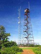 Photo of two towers that appear side by side. The tower on the left is shorter and has several discs on its side. The tower on the right has zig zag stairs going up the middle with a hut on top. Green trees appear on the right foreground with a blue sky in the background.