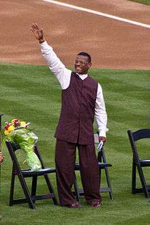 A man in brown pants, a brown vest, and white shirt raises his right hand. He is standing on grass in front of three chairs, one with a bouquet of flowers on it.